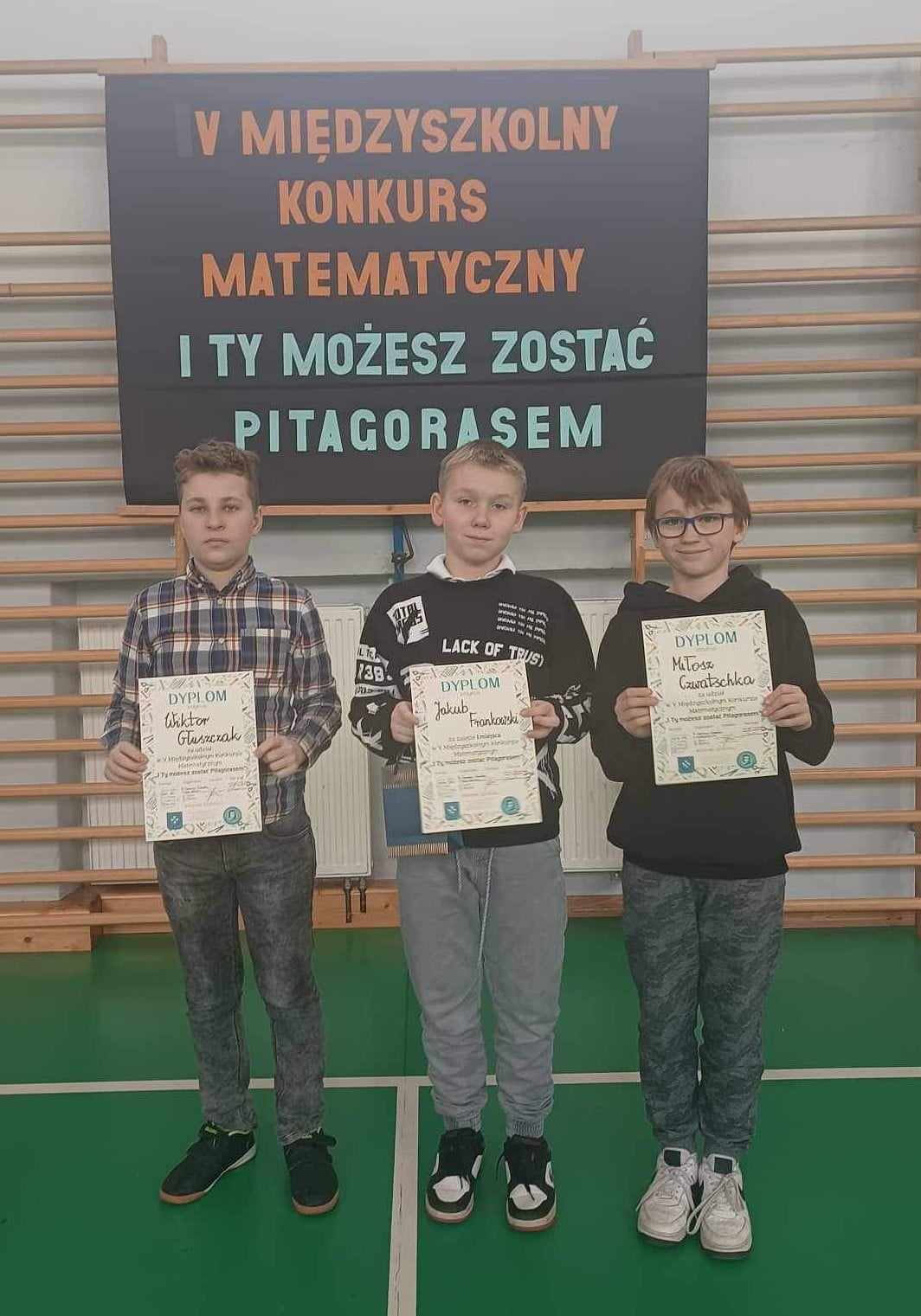 3 chłopców stoi i trzyma w rękach dyplomy. Z tyłu tablica z napisem: V Miedzyszkolny Konkurs Matematyczny I TY MOŻESZ ZOSATAĆ  PITAGORASEM.